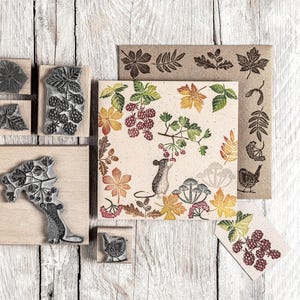 May include: A selection of wooden stamps with botanical designs, including leaves, berries, and a mouse. The stamps are displayed with a printed card featuring a mouse and colourful autumn foliage. The colour palette includes shades of brown, green, yellow, and red.