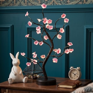 Può includere: Una lampada decorativa a forma di albero di fiori di ciliegio con luci a forma di fiori rosa. La lampada ha un tronco e rami marrone scuro e poggia su una base rotonda nera. Nelle vicinanze si trovano una statuina di coniglio in ceramica bianca e una piccola sveglia.