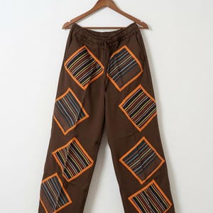 May include: Brown trousers featuring an elastic waistband with a drawstring and multiple diamond-shaped patches. Each patch displays a multicoloured striped pattern with thin orange borders, distributed across the front of the garment.