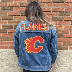 Puede incluir: Chaqueta vaquera azul medio con la palabra "FLAMES" en letras rojas y doradas en los hombros. Debajo de las letras hay un logotipo "C" rojo y dorado con un diseño de llama. La chaqueta es azul medio.