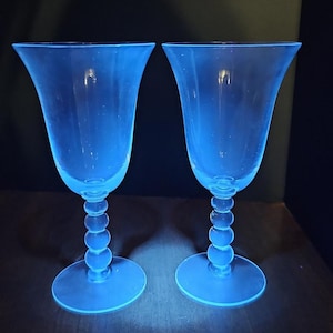 May include: Two elegant, blue-tinted wine glasses. Each glass features a flared bowl, a stem composed of stacked spheres, and a circular base. The glasses are positioned side-by-side, suggesting a set, and are illuminated against a dark background.