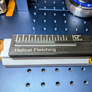 Op de afbeelding: Een zwart en grijs helical fletching-gereedschap met een liniaal en de tekst "Helical Fletching". Het gereedschap rust op een wit en grijze basis met de tekst "U-VII KARMAN". De achtergrond is een blauw oppervlak.