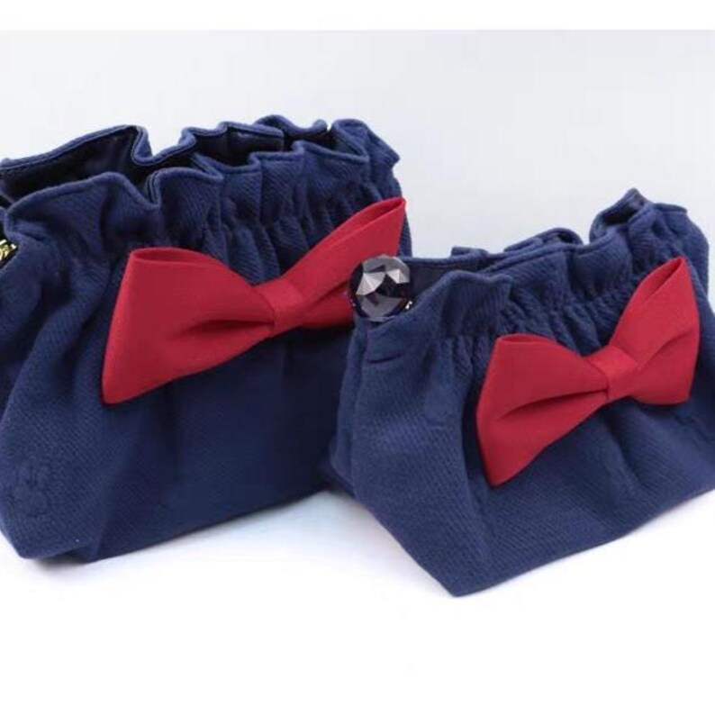 Puede incluir: Dos bolsas de tela azul marino con un borde superior fruncido y con volantes. Cada bolsa presenta un gran lazo rojo s&oacute;lido sujeto en la parte delantera. La bolsa m&aacute;s peque&ntilde;a de la derecha incluye una cuenta decorativa oscura facetada.
