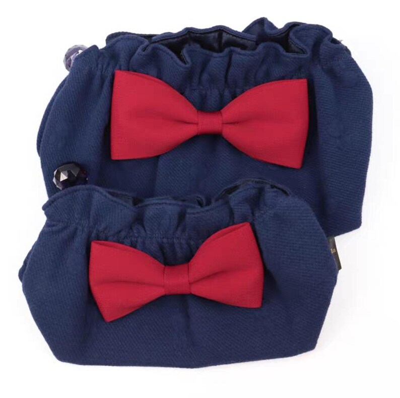 Puede incluir: Dos bolsas de tela azul marino con aberturas fruncidas y un lazo rojo oscuro sujeto en la parte frontal de cada una. Las bolsas presentan un material texturizado y un peque&ntilde;o detalle de cuenta negra en el lateral de la bolsa m&aacute;s grande.