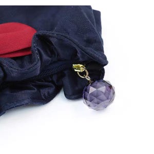 Puede incluir: Un estuche de tela azul marino oscuro con un borde fruncido y volante, y una pieza de acento en rojo intenso. El cierre de cremallera tiene un anillo de metal dorado con un dije de cuenta esf&eacute;rica, facetada y transl&uacute;cida de color morado.