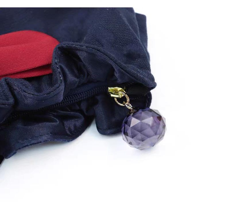 Puede incluir: Un estuche de tela azul marino oscuro con un borde fruncido y volante, y una pieza de acento en rojo intenso. El cierre de cremallera tiene un anillo de metal dorado con un dije de cuenta esf&eacute;rica, facetada y transl&uacute;cida de color morado.