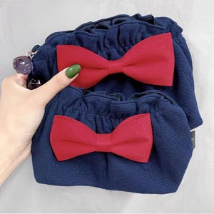 Puede incluir: Un conjunto de dos bolsos de tela azul marino con superficie texturizada, cada uno con un lazo rojo prominente en la parte delantera. Los bolsos tienen un borde superior fruncido y con volantes, y un abalorio morado facetado en la cremallera.