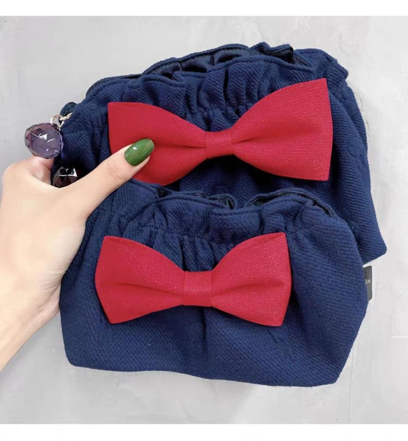 Puede incluir: Un conjunto de dos bolsos de tela azul marino con superficie texturizada, cada uno con un lazo rojo prominente en la parte delantera. Los bolsos tienen un borde superior fruncido y con volantes, y un abalorio morado facetado en la cremallera.