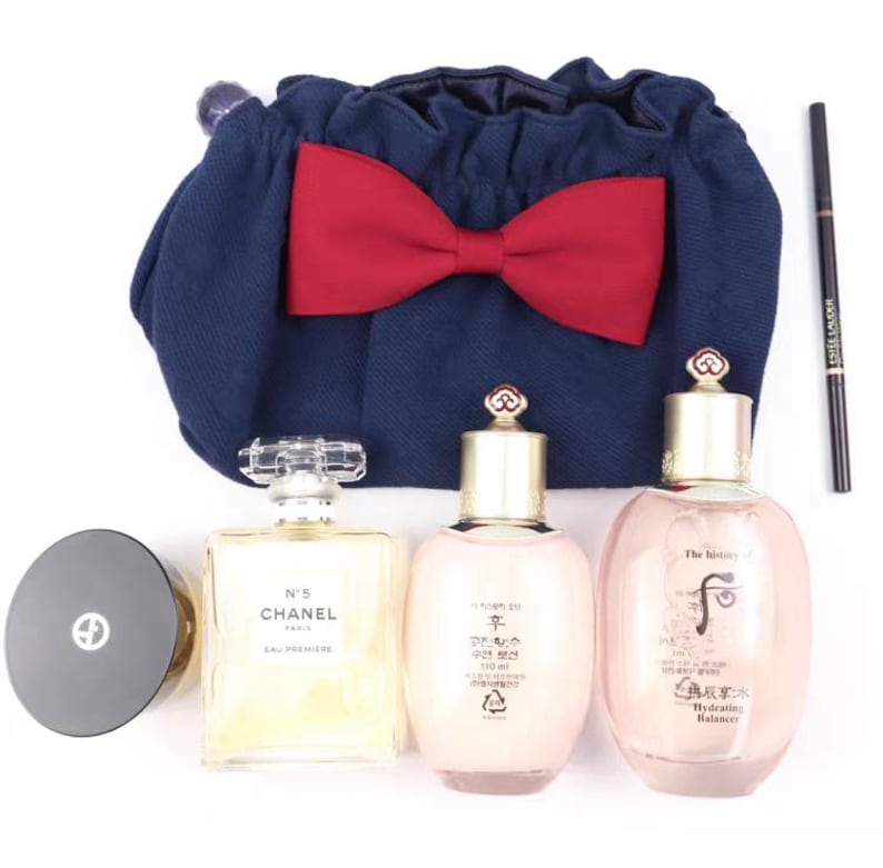 Puede incluir: Una bolsa azul marino con cord&oacute;n y un lazo rojo grande, un recipiente circular negro, un frasco de perfume rectangular Chanel No. 5 Eau Premiere, dos botellas de cuidado facial rosa claro The History of Whoo y un delineador negro Estee Lauder.