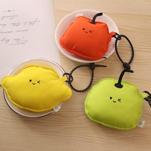May include: Three fruit-shaped reusable shopping bags in orange, yellow, and green. Each bag has a smiling face and a black stem. They are on clear plates.