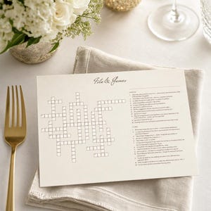 May include: A cream-colored crossword puzzle with the names "Isla & James" printed at the top. The puzzle is on a beige linen napkin, next to a gold fork and a vase of white flowers. A glass of water is in the background.
