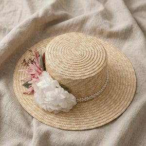 May include: A straw boater hat with a white flower and pink and green floral accents. The hat features a pearl band around the crown and a wide brim. The hat is a light tan color.