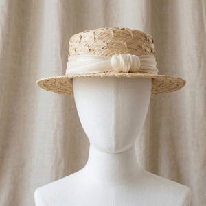 May include: A straw boater hat with a cream-colored ribbon and a decorative flower. The hat has a flat crown and brim, and is displayed on a white mannequin head. The hat is made of woven straw.