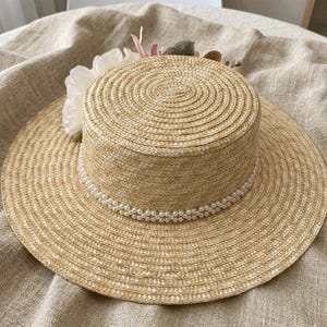 May include: A straw hat with a wide brim and a cylindrical crown. The hat is decorated with a string of faux pearls around the base of the crown and a cluster of white and pink flowers. The hat is made of woven straw.