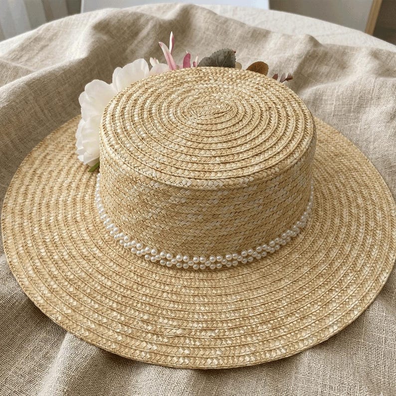 May include: A straw hat with a wide brim and a cylindrical crown. The hat is decorated with a string of faux pearls around the base of the crown and a cluster of white and pink flowers. The hat is made of woven straw.