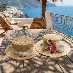 May include: Two straw hats sit on a wooden table. One hat has a white ribbon and a decorative knot. The other hat is adorned with pink and white flowers. The background shows a scenic view of the ocean and mountains.