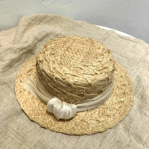 May include: A natural straw hat with a flat crown and wide brim. A cream-colored ribbon is wrapped around the crown and tied in a bow. The hat is made of woven straw and has a classic, timeless design.