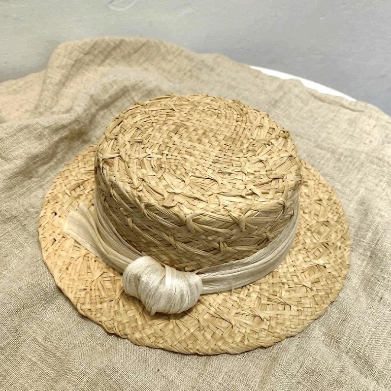 May include: A natural straw hat with a flat crown and wide brim. A cream-colored ribbon is wrapped around the crown and tied in a bow. The hat is made of woven straw and has a classic, timeless design.