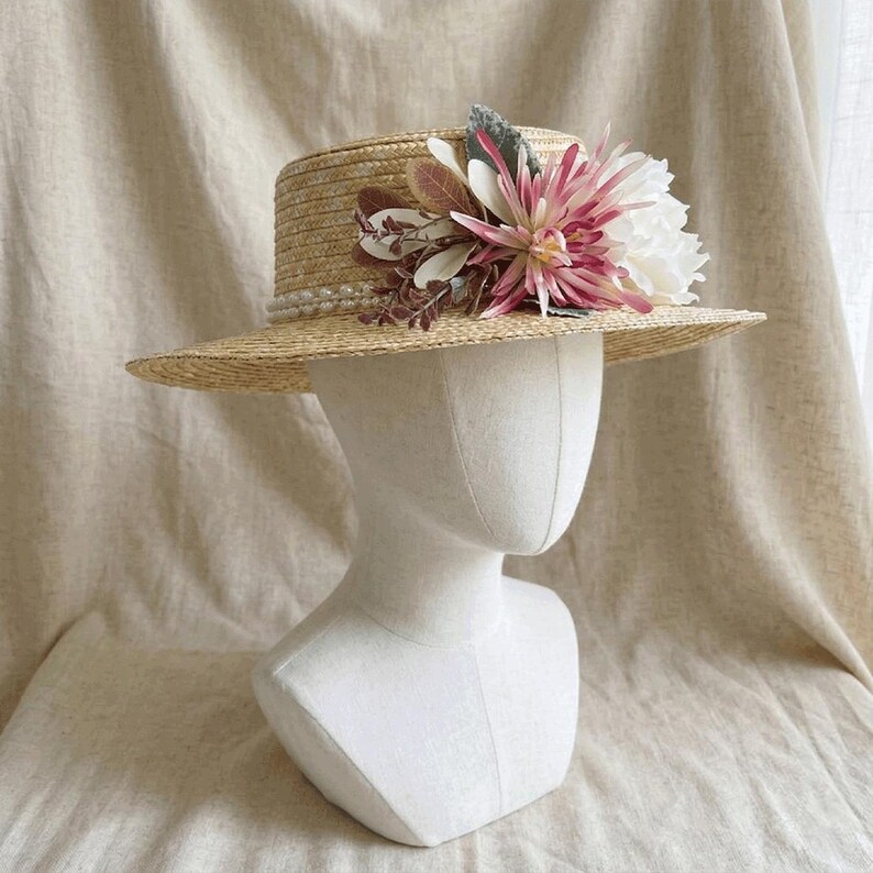 May include: A straw boater hat with a flat crown and brim, adorned with a cluster of faux flowers in shades of pink, white, and green. A string of pearls encircles the crown. The hat is displayed on a white mannequin head.