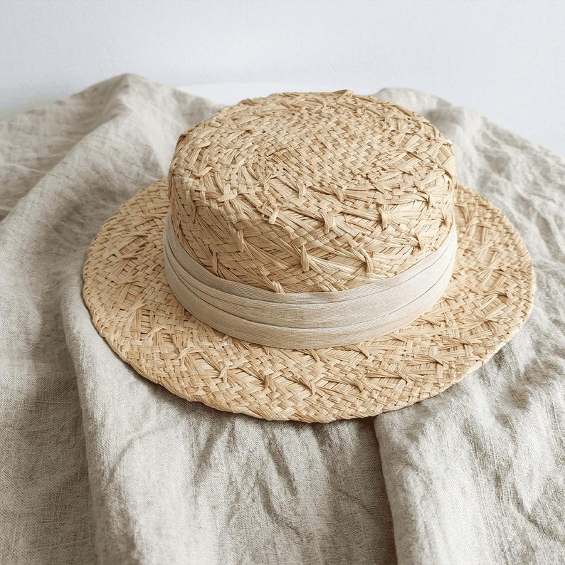 May include: A natural straw boater hat with a wide brim and a beige band. The hat is made of woven straw and has a flat top. The hat is sitting on a beige linen fabric.