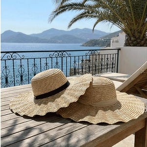 Può includere: Due cappelli di paglia da sole con ampie falde appoggiati su una superficie di legno. Un cappello ha un nastro nero, l'altro un nastro beige. I cappelli sono davanti a una vista panoramica sul mare e sulle montagne.