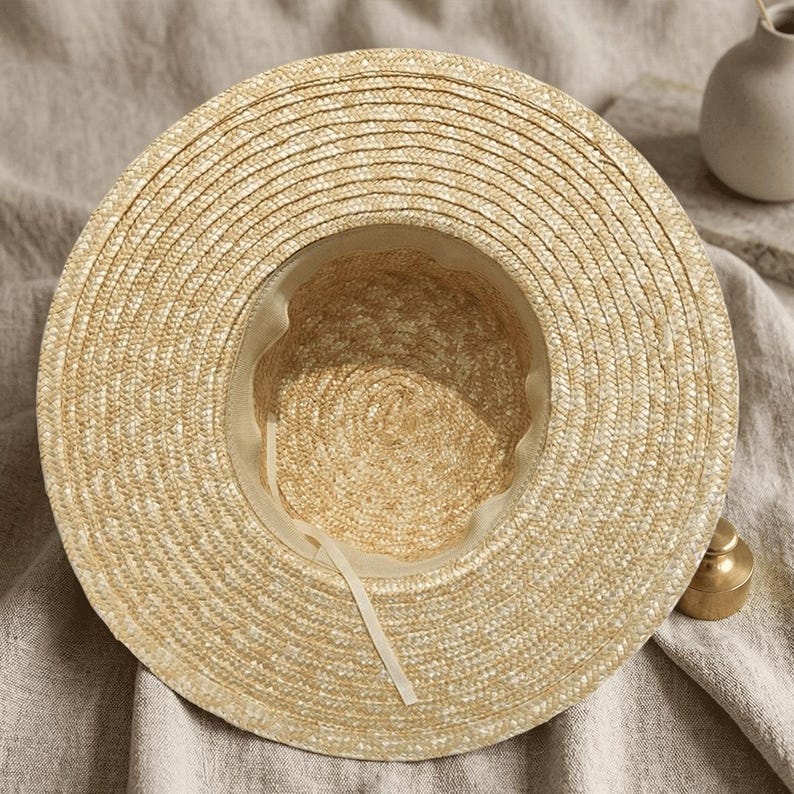 May include: A wide-brimmed straw hat with a light beige color. The hat features a circular design with a woven texture and a cream-colored interior lining. A cream-colored ribbon is attached inside the hat.