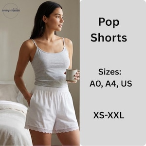 Puede incluir: Una mujer lleva un top de tirantes gris claro y unos shorts blancos con ribete de encaje. Los shorts tienen bolsillos. El texto "Pop Shorts" y las opciones de tallas son visibles en la imagen.