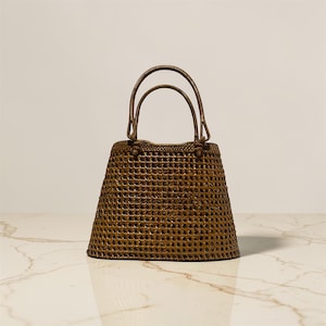 May include: A brown woven handbag with two arched handles. The bag has a rectangular base that widens towards the top, with a textured, open weave pattern. The handles are attached to the top of the bag.