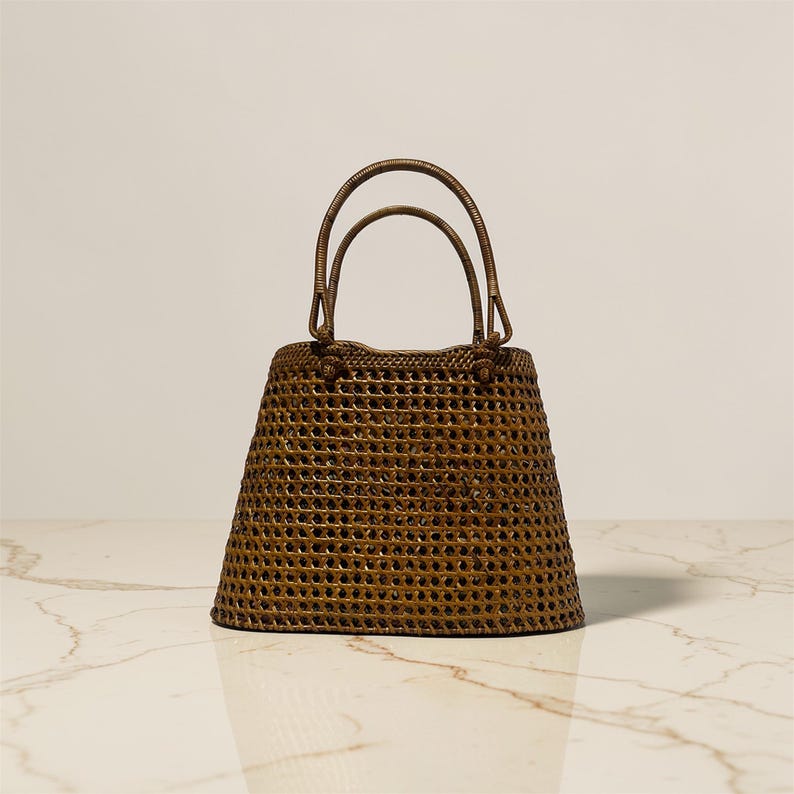 May include: A brown woven handbag with two arched handles. The bag has a rectangular base that widens towards the top, with a textured, open weave pattern. The handles are attached to the top of the bag.