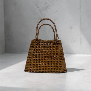 May include: A brown rattan handbag with a woven design and two arched handles. The bag has a trapezoidal shape with a wide, open top. The bag is made of rattan and is sitting on a white surface.