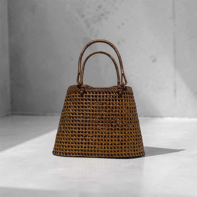 May include: A brown rattan handbag with a woven design and two arched handles. The bag has a trapezoidal shape with a wide, open top. The bag is made of rattan and is sitting on a white surface.