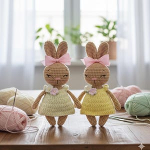 May include: Two crocheted bunny dolls. Each has a light brown body, a pink bow, and a yellow dress with a flower. The dolls are holding hands. Yarn and knitting needles are in the background, on a wooden table.