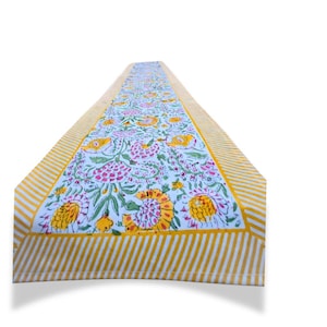May include: A rectangular table runner with a floral pattern in pink, yellow, and green on a white background. The edges feature a yellow and white striped border. This decorative runner is suitable for dining tables.
