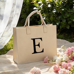 Puede incluir: Un bolso tote beige con una gran letra "E" negra impresa en la parte delantera. El bolso tiene dos asas y está colocado sobre una manta con flores rosas y blancas. El fondo es un área verde y frondosa.