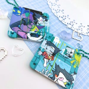 May include: Two square teal plastic pendants featuring printed illustrations of a cartoon character with a long nose, dark hair, and colorful clothing. Each pendant is attached to a green ball chain and displays a unique graphic design.
