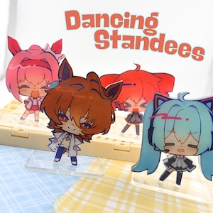 May include: Four acrylic standees featuring chibi-style anime characters with colorful hair and outfits. The characters have distinct features including cat ears and headsets. The text "Dancing Standees" is printed in orange above the figures.