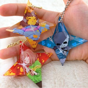 Puede incluir: Tres llaveros de acrílico en forma de estrella con personajes de estilo anime. Un dije de tonos amarillos muestra un personaje con camisa a rayas azules y moradas, uno azul muestra un esqueleto y uno rojo muestra un personaje con un cuchillo.