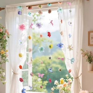 May include: A decorative hanging string curtain featuring an assortment of colorful artificial flowers and butterflies in shades of pink, blue, yellow, red, and white, suspended vertically in front of a window with sheer white curtains.