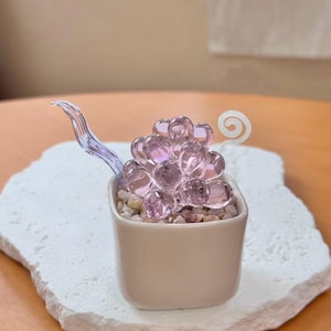 May include: A decorative glass sculpture of a bunch of grapes in a small, square, off-white ceramic pot. The grapes are made of translucent pink glass and sit atop small pebbles. The pot is on a white, textured, irregularly shaped base.