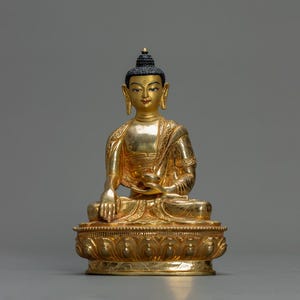 May include: A golden Buddha statue in a seated position, with intricate detailing on the robes and base. The statue has a serene expression, with a black bun on top of the head. The statue is made of a shiny, reflective material.