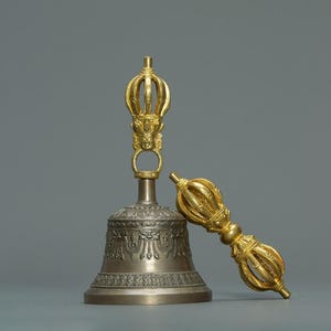 Puede incluir: Un juego de campana y dorje budista de plata y oro. La campana tiene intrincadas tallas y un asa dorada con un diseño de corona detallado. El dorje, o rayo, es dorado y descansa junto a la campana. El fondo es gris liso.