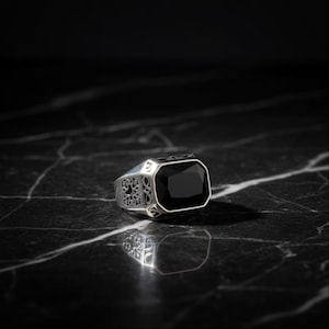 以下が含まれることがあります： 大きな長方形の黒い宝石が付いたシルバーリング。リングの側面には複雑なフィリグリーのデザインがあり、研磨仕上げが施されています。リングは黒い大理石の表面に表示されています。