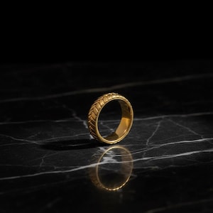 Puede incluir: Un anillo dorado con un diseño texturizado y tejido. El anillo se encuentra sobre una superficie oscura, similar al mármol, con su reflejo debajo. El diseño del anillo presenta una serie de líneas anguladas repetidas, creando una apariencia trenzada.