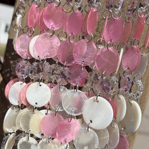 Puede incluir: Un carillón decorativo con discos de concha iridiscentes rosas, blancos y transparentes y detalles de cristal. Los discos están conectados por cadenas plateadas y suspendidos de un pasador de madera.