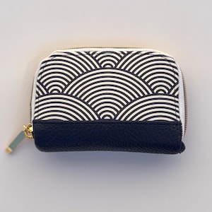 May include: A small, zipped wallet. The top half features a black and white wave pattern, while the bottom is navy blue leather. The zip is gold-coloured, and the pull tab is a light blue-green colour.
