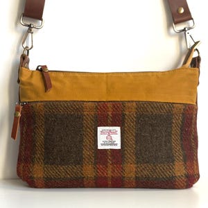 Peut inclure: Sac bandoulière à motif écossais marron, rouge et or. La partie supérieure est jaune moutarde. Il possède une bandoulière et des tirettes de fermeture éclair en cuir marron. Une petite étiquette blanche est visible.