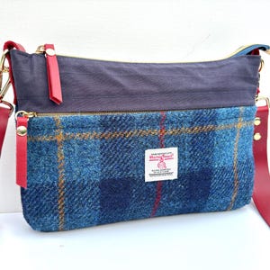 Peut inclure: Un sac bandoulière avec un panneau avant en laine Harris Tweed à carreaux bleus portant une étiquette blanche, une section supérieure gris foncé et une bandoulière en cuir rouge avec des ferrures dorées et deux fermetures éclair rouges.