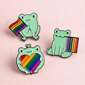 Könnte beinhalten: Drei Emaille-Anstecknadeln mit hellgrünen Fröschen. Ein Frosch hält eine Regenbogenflagge, ein zweiter sitzt neben einer vertikalen Regenbogenflagge und der dritte hält ein Regenbogenherz. Alle Anstecker haben schwarze Konturen und eine glatte Oberfläche.