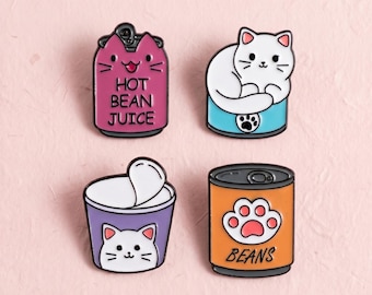 Könnte beinhalten: Vier Emaille-Anstecknadeln mit Katzenmotiven. Eine ist eine rosa Dose mit der Aufschrift HOT BEAN JUICE, eine zeigt eine weiße Katze auf einer blauen Dose, eine ist ein violetter Becher mit Katzengesicht und eine orange Dose mit Pfotenabdruck und BEANS.