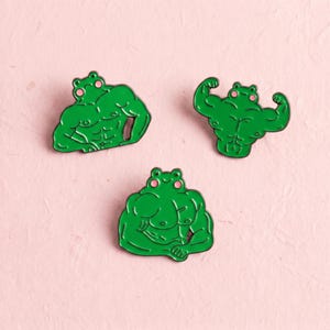 May include: Three green enamel pins featuring muscular frog characters with pink cheeks. Each pin depicts a different pose of a frog with defined abdominal and arm muscles, rendered in a cartoon style with black outlines and vibrant green enamel fill.